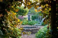 View through arch of Vitis vinifera 'Purpurea' to formal walled garden with brick path, well, antique water pump and herbaceous borders with Malus - Apple tree. Parham, Sussex