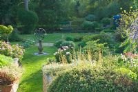 The upper garden has a sundial at its centre, and shaped beds full of herbs, herbaceous perennials including Peonia and interesting shrubs with mill pond seen beyond. Mill House, Netherbury, Dorset, UK