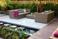 Small garden with wicker sofas on decked and paved patio, backed by Fargesia murielae - Bamboo hedge
 