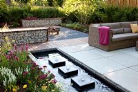 Small garden with wicker sofas on decked and paved patio, backed by Fargesia murielae - Bamboo hedge. Rectangular pond with row of square water features 