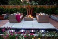 Small garden lit up at night, with wicker sofas on decked and paved patio, backed by Fargesia murielae - Bamboo hedge. Rectangular pond with row of square water features and lights  