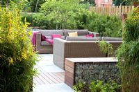 Small garden with wicker sofas and Gabion benches on decked and paved patio, backed by Fargesia murielae - Bamboo hedges.