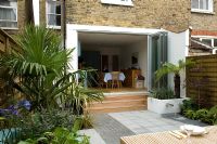 Small urban garden with with paved patio area and sliding French door, Trachycarpus to left and small water feature with spout