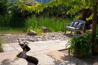 A meandering canal leads through a flagstone paved area to a natural swimming pool that is formed with pebbles and gravel, with a wooden bench with linen cushions. Planting includes Agapanthus, Centranthus ruber 'Coccineus', Gleditsia triacanthos 'Sunburst', Ranunculus lingua, Scirpus and Typha 