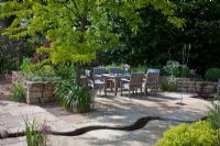 Raised dry stone wall bed, a rest area with wooden furniture and a meandering canal through the flagstone paving. Planting includes Agapanthus, Allium christophii, Centranthus ruber 'Coccineus', Gleditsia triacanthos 'Sunburst' and Tanacetum parthenium 