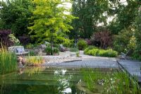 Natural swimming pool with pebble 'beach zone', meandering canal in flagstone paving, different rest areas, perennials and shrubs against the background big trees. Planting includes Agapanthus, Alchemilla mollis, Cotinus coggygria 'Royal Purple', Gleditsia triacanthos 'Sunburst', Physocarpus opulifolius 'Diabolo', Ranunculus lingua and Typha 