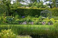 View over a natural swimming pool to a pavillon, backed with a hedge. The slope is secured by lime sand boulders, planting includes Alchemilla mollis, Buxus, Carpinus betulus, Fagus, Geranium, Hosta, Larix, Lupinus and Salix

