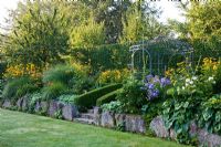 Lime sand boulders secure a slope with perennials, stone steps lead to an iron cast pavillon backed by a hedge. Planting includes Alchemilla mollis, Hostas, Buxus, Carpinus betulus, Fagus, Heliopsis, Hosta, Miscanthus sinensis, Phlox paniculata and Rudbeckia fulgida
