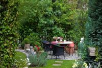 Seating area with a decorated table, black furniture and containers on paved circle. Other planting includes Chamaecyparis lawsoniana, Hedera helix and Lavandula