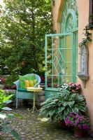 Turquoise wicker chair and yellow bistro table on paved patio with plants in terracotta pots next to classical styled glass door - Hosta, Ilex aquifolium, Impatiens and Oxalis