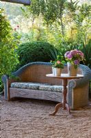 Wicker sofa and a table with plants in tin pots on a gravel area. In the background a border which contains Box sphere, Hydrangea macrophylla, Olea europaea, Taxus baccata, Wisteria and Yucca