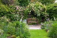 Wooden bench on paved area under a rose arbour in a romantic garden with roses and perennials - Rosa 'Ghislaine de Féligonde' and 'Rosenprofessor Sieber', Aquilegia, Campanula persicifolia, Digitalis and Matricaria