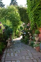 Stone busts on plinths and an angel line a pathway paved with flagstone and granite. Planting includes Aristolochia macrophylla, Clematis and roses