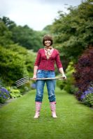 Woman wearing blue jeans and wellies standing on a lawned path carrying a garden fork 