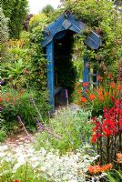 Wooden archway surrounded by a mass of colourful, flowering plants including Crocosmias, Salvia leucantha, Lobelia cardinalis 'Queen Victoria', Helichrysum and Clematis. Poppy Cottage Garden, Roseland Peninsula, Cornwall, UK