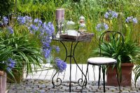 A metal chair and bistro table with chandeliers in front of natural swimming pool. Planting of Agapanthus africanus 'Blue Star'