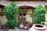 Display of terracotta and decorative metal objects on a bistro table in a gangway. Plants in containers are Laurus nobilis