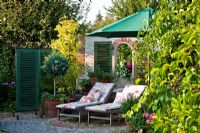 Next to backdrop with green shutters two loungers beneath green parasol on a granite paved rest area surrounded by Juniperus 