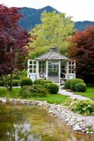 White painted wooden pavilion with a group of box spheres, mature trees and a natural swimming pool. Planting includes Acer negundo 'Variegatum', Acer palmatum, Acer palmatum 'Dissectum', Buxus and Hosta