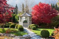 A flagged path and white painted wooden pavilion next to Acer palmatum - Japanese Maples in autumn colours. Buxus - Box balls