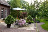 A flagstone paved terrace with garden furniture and a parasol. Planting includes - Acer palmatum 'Atropurpureum', Allium nigrum, Camellia japonica, Geranium 'Sirak', Heuchera micrantha 'Palace Purple' and Prunus domestica