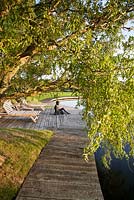 Boardwalk, chairs and sculpture alongside landscaped lake with overhanging Willow in late summer. Statue created by Brian Alabaster
