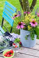 On a wooden table a vase with coneflower, Ladys Mantle, Veronica and grapes. Also on the table is a gardening book, gardening gloves, hand fork a bowl of strawberries and a glass of water. In the background a colourful turquoise garden chair.