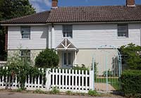 Weather boarded cottage with white picket fence and painted antique wrought iron garden gate.