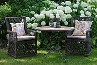 Two wicker armchairs with embroidered patchwork cushions and a bistro table with candle holders against a backdrop of white flowering Hydrangea arborescens 'Annabelle'