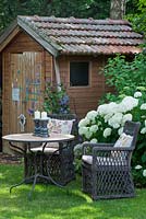 In a shaded garden next to summerhouse, a rest area with two wicker armchairs with cushions and a bistro table. White flowering Hydrangea arborescens 'Annabelle'
