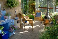 Wicker seats in covered outdoor living area with stone floor, hanging baskets, colourful containers, painted dressing table with Petunia in open drawer and chiminea