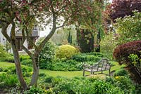 View in spring of island bed beside house with garden seat beneath Malus floribunda in bud -Glen Chantry, Essex