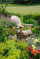 Adirondack garden chair surrounded by Geranium psilostemon and Alchemilla mollis. Containers with Pelargoniums, Violas, Nemesias and Marguerites. Hedge with meadow in background