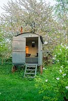 Shepherds hut in wild garden with Prunus avium, wild cherry, behind and Exochorda x macrantha 'The Bride' in foreground - The Mill House, Little Sampford, Essex