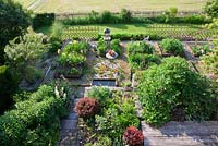 View from above onto a modern structured country garden with a small rest area, vegetable patches and a wooden terrace. Plants include Berberis thunbergibi 'Atropurpurea', Dictamnus albus and Paeonia