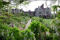 Gravetye Manor in summer. View to the house through Wisteria floribunda 'Alba', with Digitalis 'Suttons Apricot' in front of Euonymus 'Silver Queen' and Lychnis flos-cuculi 'Jenny', Angelica  and Papaver commutatum.