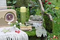 A rustic, moss covered bench with thermos flask, lunchbox, woollen throw, sock knitting, vase of snowberries, Bush radio and Observer books