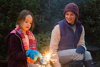 Mother and daughter lighting sparklers by a firepit in an autumnal back garden.