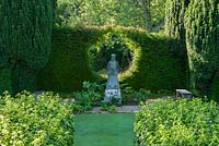 Statue of oriental priest is framed by a circle cut into a yew hedge that supports a honeysuckle, viewed from top of steps leading through wall clothed in Hydrangea petiolaris. Melplash Court, Bridport, Dorset, UK