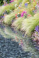 Stipa tenuissima and Ajuga, Zinnia and Geum planted on a bank bordering the waters edge. The Rush of Nature exhibition garden designed by Marc Quinn.
