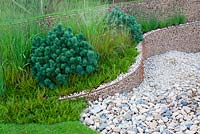 Pebble stone pathway and walls made of metal mesh and infilled with gravel with border between planted with grasses Calluna and conifer in the Natuarl Forces garden at RHS Tatton Flower Show 2013