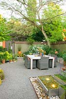 Summer outdoor living space with dining table and chairs lighted fountain, and enclosed with wooden fence. Albizia julibrissin - Persian Silk Tree syn. Mimosa Tree.