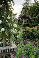 View of floral scene of roses on a wooden obelisk and an arch with geranium psilostemon. Stone water feature glimpsed in next garden room.  Seend, Wiltshire