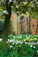 View across spring border to ornamental wrought iron gate brick wall and obelisk. Hertfordshire Heydonbury 
