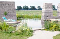 Towers made of recycled paving stones in a swimming pond with relaxing chair on a deck of concrete. Wildflowers like Senecio jacobaea and Anthemis arvensis growing in the landscape.