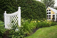 White wooden fence with trellis frames next to a flowerbed of yellow daylilies (Hemerocallis) and an artist's workshop surrounded by a cedar hedge (Thuja occidentalis)  - Il Etait Une Fois garden, Monteregie, Quebec, Canada. 