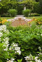 White Hydrangea paniculata 'Quick Fire' with orange and red daylilies 'Hemerocallis' and the back of a brown metal lattice garden bench. Miscanthus sinensis 'Berlin' ornamental grass plants in the background - Il Etait Une Fois, Monteregie, Quebec, Canada. 
