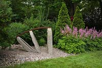 Concrete and iron sculpture next to a patch of pink Astilbe 'Ostrich Plume' flowers in a border - Il Etait Une Fois, Quebec, Canada. 
