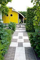 Black and white pavement in the entrance. Bright yellow shed with roundshaped windows. Rudbeckia fulgida Goldsturm in the border.