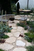 Hambrooks Halo garden  Mediterranean Greek island style. Rough-hewn stone paving with circular conversation seating with fire pit and table and chairs. Coastal seaside planting with verbasum. Sails in background. Designer: Stuart Charles Towner Silver-Gilt award.  